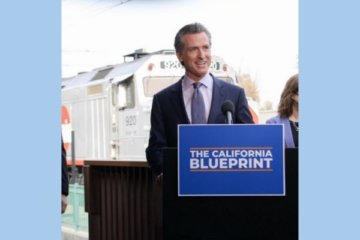 Image is a male California Governor standing in front of a podium speaking at a train station. There is a train traveling behind the man. The podium has a microphone and a sign that reads “The California Blueprint.” 