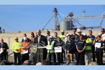 Press conference with a man speaking at a podium with a little over a dozen people standing in the background. Conference is outside on a sunny day. People in background are wearing solid blue shirts or construction hats and safety vests.
