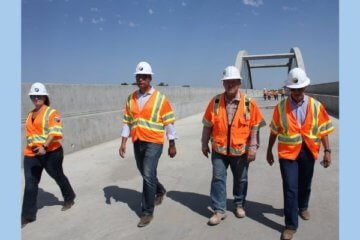 Four people walking on a viaduct structure during the day. One woman and three men are in the photo all wearing jeans, security vests and hardhat construction helmets. 