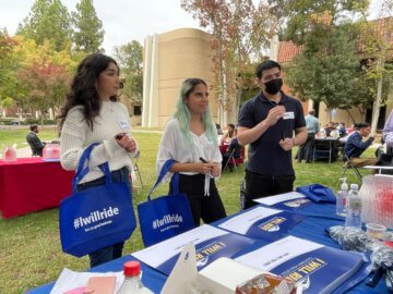 three students standing in front of a table at a networking event. Students are holding bags that read I Will Ride. 