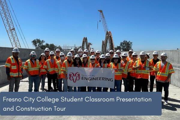 Under clear blue skies, a group of 30 or so students and professors stand holding a Fresno City College banner while on a construction site. All those photographed are in safety vests and construction hard hats. 