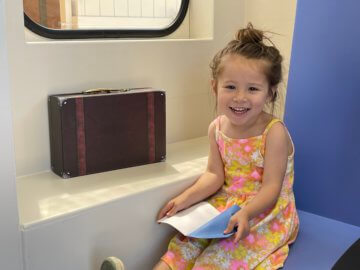 Young child sits in train exhibit seat holding a booklet and smiling. 