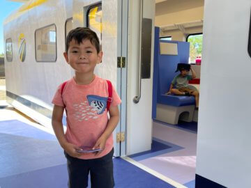 Young child stands smiling in front of train exhibit. 