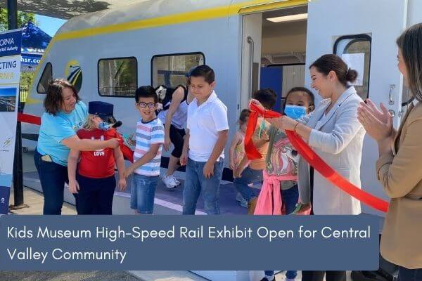 Kids and adults stand behind a red ribbon ready to cut the ribbon to show the opening of a new museum exhibit. Behind the people is a large train in a kid’s museum exhibit where kids can play on the train. 