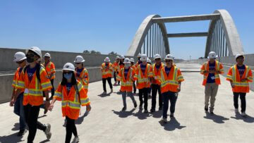 Students in construction safety equipment walk across a large bridge structure.