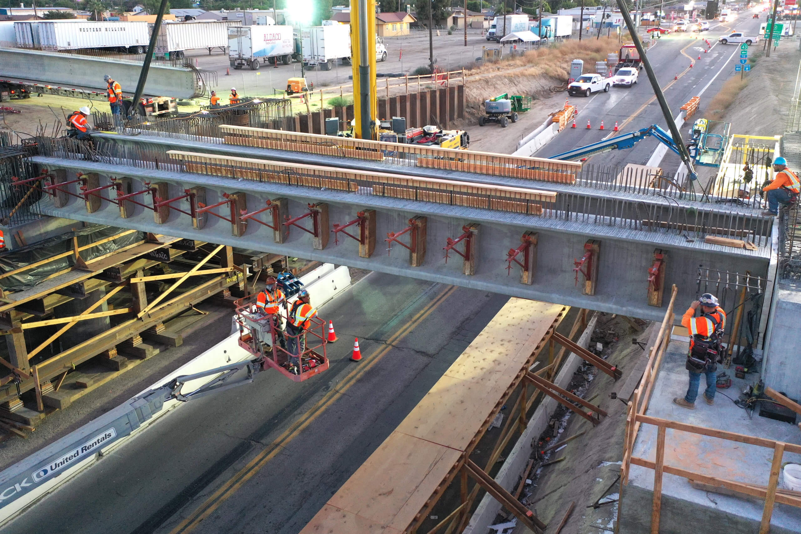A shot from above, showing a concrete girder being placed on columns and abutments by a crane over state route 46 at dusk. Other heavy machinery and workers and vehicles also visible.
