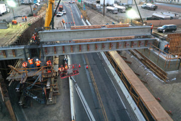 A shot from above, showing a concrete girder being placed on columns and abutments by a crane over state route 46 at dusk. Other heavy machinery and workers and vehicles also visible.