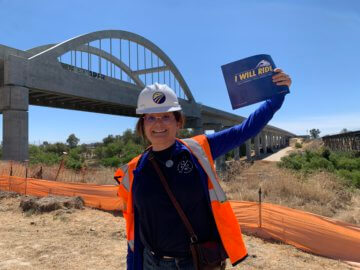 Teacher in construction safety equipment holding a sign and smiling in front of a large bridge structure with arches. 
