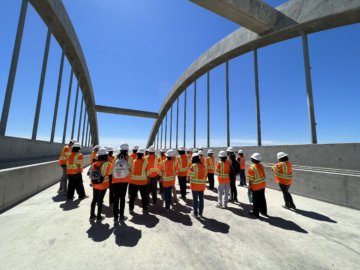 Students in protective construction gear stand on top of a bridge structure in between large arch structure. 