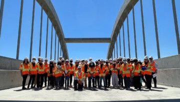 Photo of 40 or so people on a bridge structure. The students face the camera and in the background is a large structural arch. Photo was taken in the day and there are clear skies and the sun is very bright. 