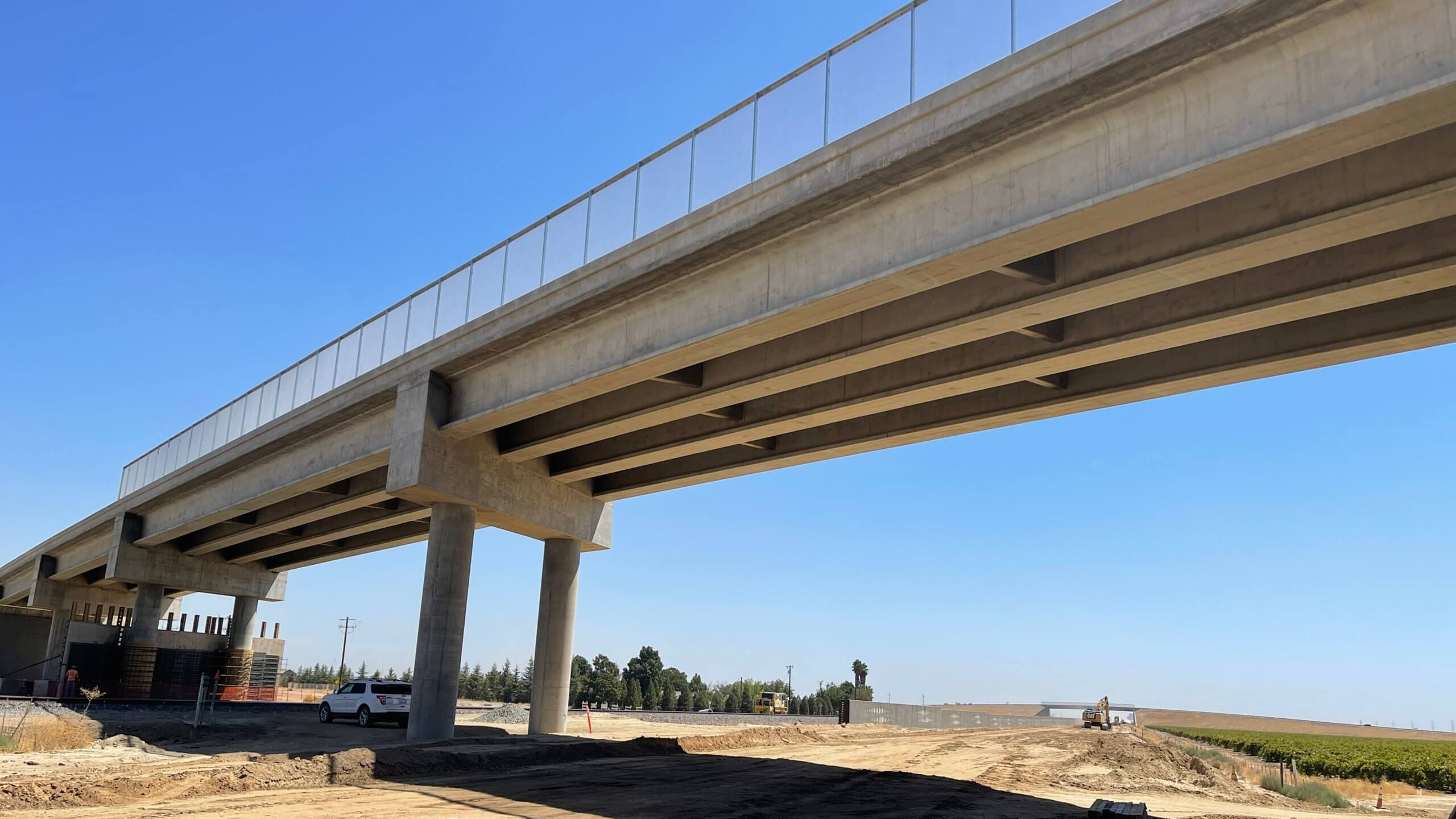 Underneath the newly compeleted and opened Avenue 15 1/2 grade separation, in the right of way where tracks will be laid. Another grade separation is visible on the horizon. Bright blue sky.