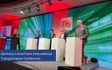 Four-person panel and a moderator stand on a stage. The five people on stage stand in front of a podium. There is an extravagant digital background behind the speakers and a logo that reads DB. Title included in the graphic that reads Germany's InnoTrans International Transportation Conference. 