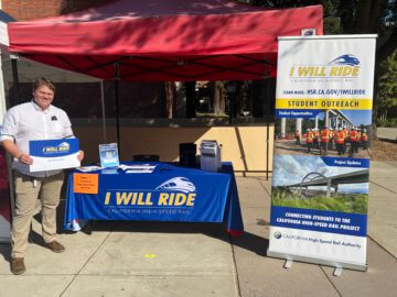 Someone holding up an I Will Ride sign next to a high-speed rail outreach table. Photo is outside on a sunny day. 