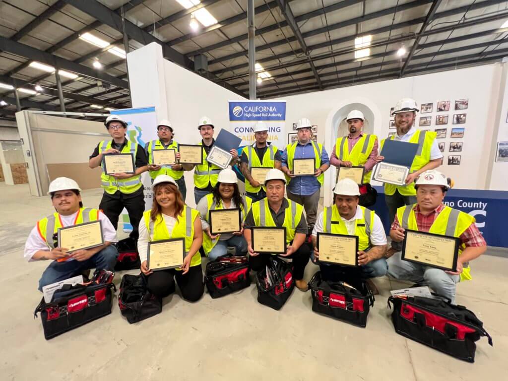 A class photo of every member of the graduating cohort, holding certificates. All are wearing personal protective equipment like high visibility vests, and helmets. Photo is inside a warehouse, in front of some of the structures they studied and built. New tool bags, given in recognition of their accomplishment, rest in front of them.