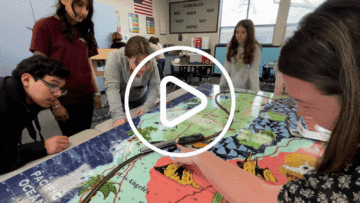 Students gather around their project on a table in a classroom. One individual points at a model freight train on tracks on top of a big, stylized map of California, spread out on the table.