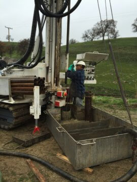 A man in a black and blue flannel shirt, green gloves, jeans, and a construction hat setting up a big white drill. The drill is mobile and has tracks underneath. In the background there are green hills, phone wires, and the day is cloudy.