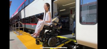 Picture of a woman using a motorized wheelchair descending off a red, grey, and white train on an accessible train bench onto a train platform. The woman is wearing a flowy white blouse and pants, with white heels and glasses. Her hair is dark and in a braid over her right shoulder.