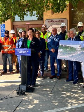 A woman in a dark pantsuit and green shirt, standing behind a podium that reads “Investing in America.” Behind the woman are six men in construction gear, including helmets and safety vests. There are also three men in suits. The entire group is under a tree in front of a building.