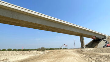 A cloudless sky is the backdrop for the Davis Avenue Overcrossing, shot from below. Dry flat earth is below, which will be the route the tracks take under Davis Avenue. A worker crane is in frame, as well as a small number of trees from surrounding lush orchards.