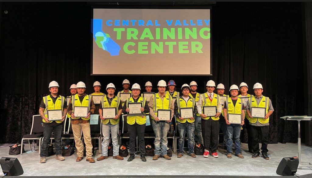 A group of people in yellow safety vests hold their diplomas in front of a screen that says Central Valley Training Center