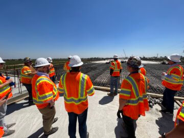 Tour goers clad in personal protective equipment survey construction, a cloudless sky overhead.