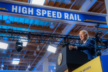 Man in a blue suit at a podium in a stadium with a banner that says “High-Speed Rail” and features a picture of two trains.