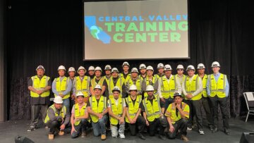 A group of people in personal protective equipment, including yellow safety vests and hard hats pose in front of a screen that says Central Valley Training Center.