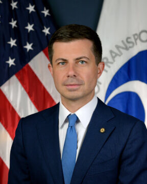 Man wearing a dark blue suit, white shirt, and blue tie in front of an American flag and a U.S. Department of Transportation flag. He has short brown hair.