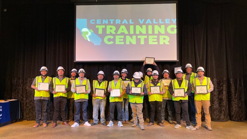 The individuals of the 12th cohort grin and stand as a group, holding their certificates, on the stage for the graduation ceremony. All are wearing personal protective equipment, such a s reflective vests, hard hats, and safety glasses. A projected screen behind them reads “Central Valley Training Center.”