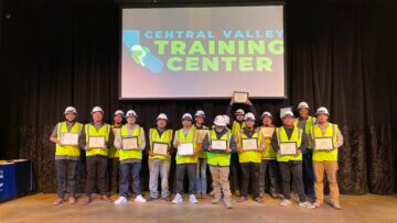 The individuals of the 12th cohort grin and stand as a group, holding their certificates, on the stage for the graduation ceremony. All are wearing personal protective equipment, such a s reflective vests, hard hats, and safety glasses. A projected screen behind them reads “Central Valley Training Center.”