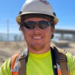 A man in a hard hat, sun glasses, and brightly colored safety gear smiles at the camera