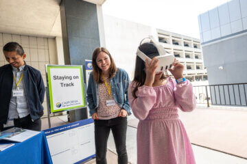 A student in a pink dress wearing a VR headset next to two HSR employees at their booth