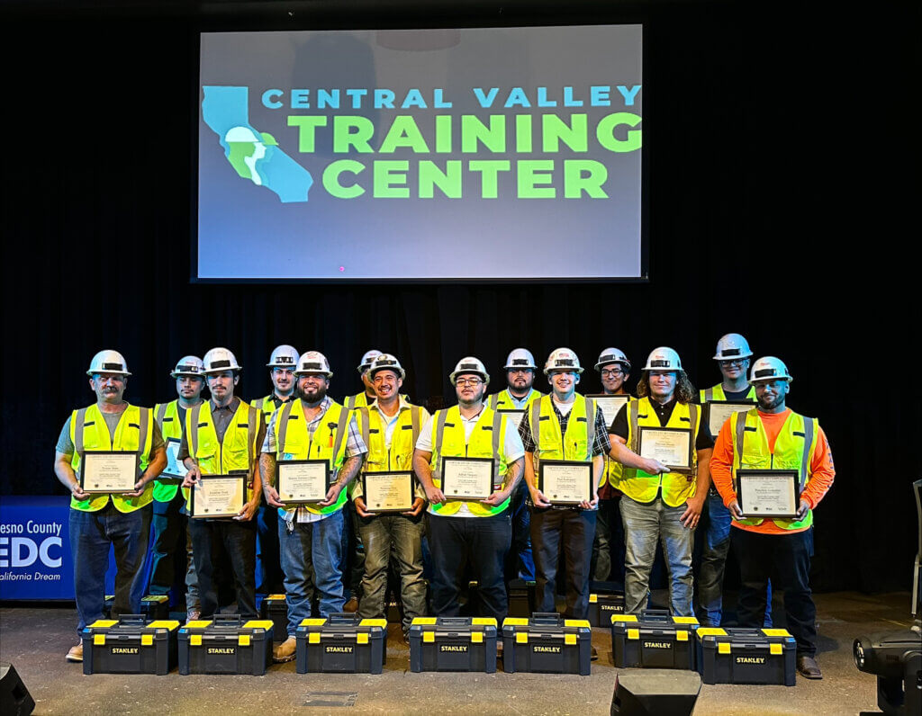 A group of people in hard hats and high-visibility vests hold framed diplomas with tool boxes at their feet. Projected above them is the Central Valley Training Program logo