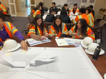 During an interactive activity, Southern California students sit at a table writing notes onto a large poster of a train interior illustration. 