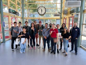 A group of 11 or so Stanford students and two professional staff members pose for a group photo at a Northern California transit station. The station is lined with glass windows and doors, providing a very open and bright space.