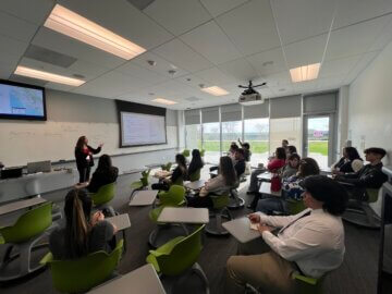 Women doing a classroom presentation in front of two dozen students or so.