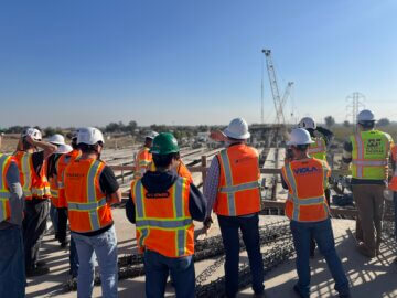 Students stand on a viaduct structure and learn about the structure while wearing safety vests and hardhats. To the horizon are two large construction cranes.