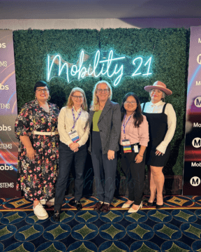 Five women in front of fake, green bush background with neon lights overhead that read “Mobility 21.”