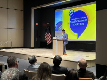 CEO Ian Choudri on stage at the California High-Speed Rail Industry Forum. He's standing at a podium in front of an audience. Behind him is a graphic image of a high-speed rail train rendering, California High-Speed Rail Authority logo, and a chat bubble that says, 