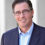 Headshot of Jeffrey Worthe. He is a wearing a suit jacket, button up shirt, and glasses. He is standing against a blurred background.