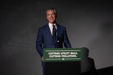 Governor Gavin Newsom in a dark blue suit, white button-up shirt, and dark blue tie. He's standing against a gray background. In front of him is a podium that says "Cutting Utility Bills. Cutting Pollution."