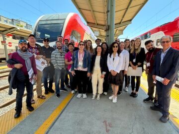 A group of Northern California Authority staff and global transportation professionals on a Caltrain platform with multiple trains in the background.