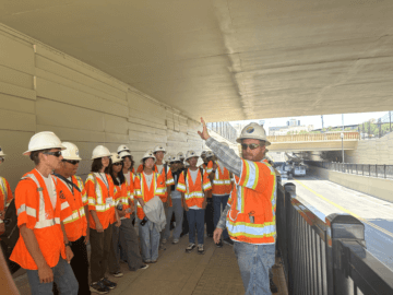 A man in an orange safety vest and hard hat talks to a group of students all wearing safety vests and hard hats. They are standing under one of the bridges of the Tulare Street Undercrossing.