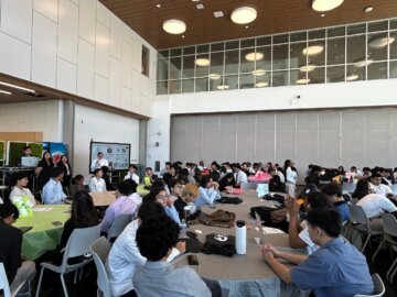 A large auditorium filled with students seated at a series of circular tables. At the front, Authority staff are giving a presentation on the I Will Ride program on a monitor.