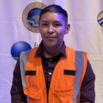 Yvette Castro, a graduate of the Central Valley Training Center, stands in front of a white backdrop featuring multiple logos. She is wearing a white hardhat and a bright orange construction safety vest, symbolizing her entry into the construction workforce.