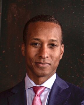 A man in a blue suit, button up shirt, and pink tie with his arms crossed. The man is standing against a dark background that looks similar to a galaxy or small drops of paint splattered on a black canvas.