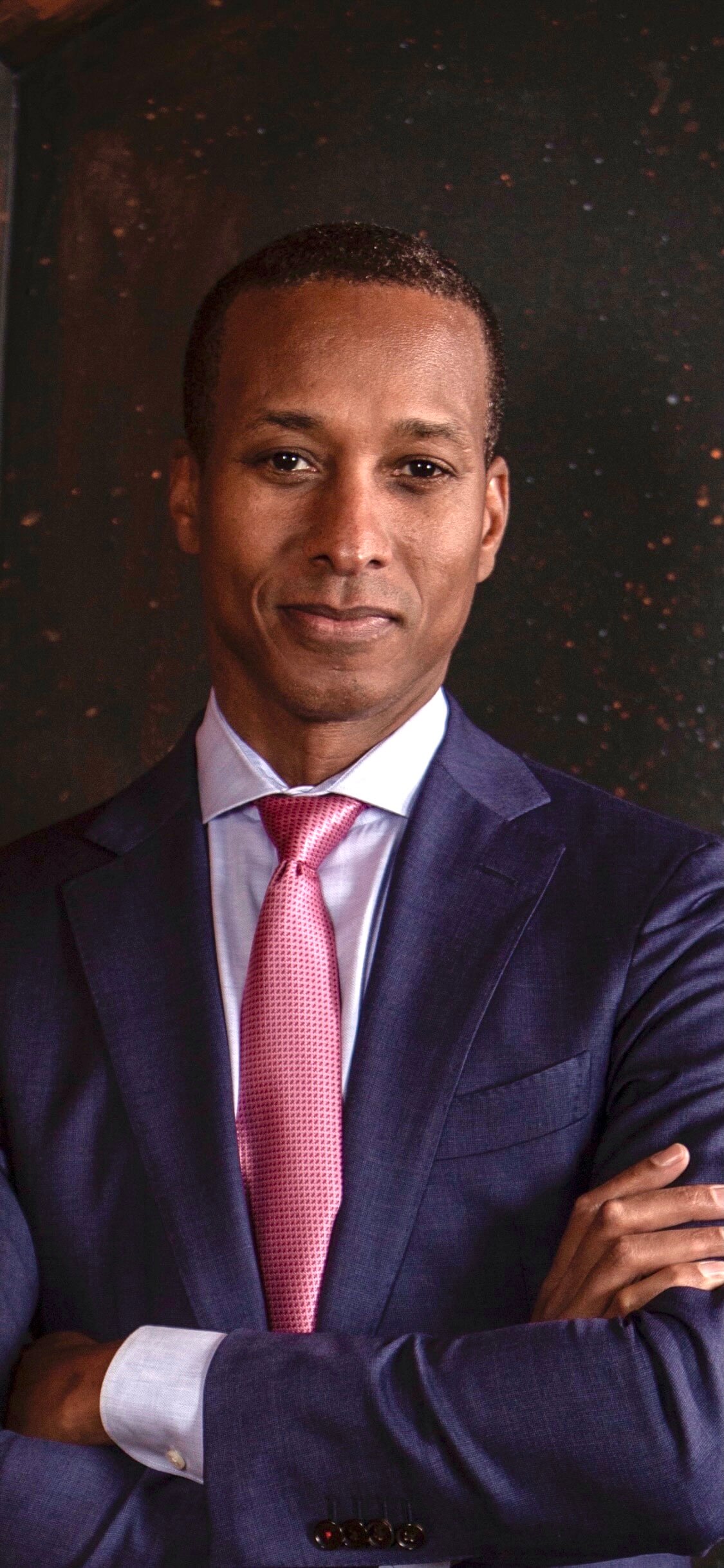 A man in a blue suit, button up shirt, and pink tie with his arms crossed. The man is standing against a dark background that looks similar to a galaxy or small drops of paint splattered on a black canvas.