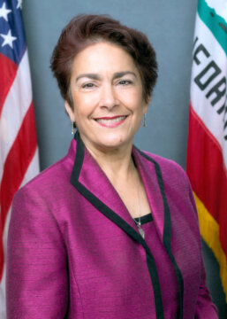 Headshot of Senator Anna Caballero. She is wearing a pink blazer and blouse and standing in front of the American and Californian flags.