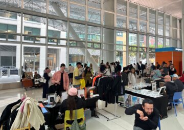 Approximately 20 members of the public circulate in the Salesforce Transit Center among several tables full of art prints and other pieces of artwork for sale.
