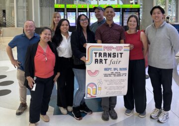 Nine staff members from the California High-Speed Rail Authority and the Transbay Joint Powers Authority in business casual attire gather at the Salesforce Transit Center behind a poster advertising a Pop-Up Transit Art Fair.